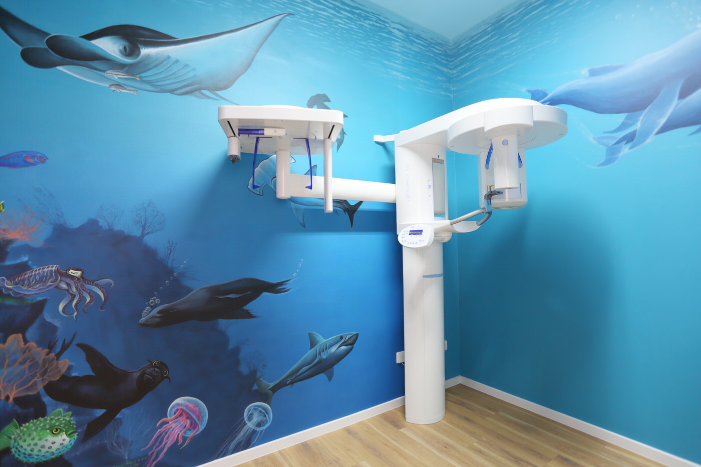 Zahnmedizinischer Röntgenraum mit kindgerechter, farbenfroher Wandgestaltung im Unterwasserstil. Die Wände zeigen Meeresbewohner wie Rochen, Haie und Quallen. Im Raum steht ein modernes 3D-Röntgengerät vor türkisblauer Kulisse.