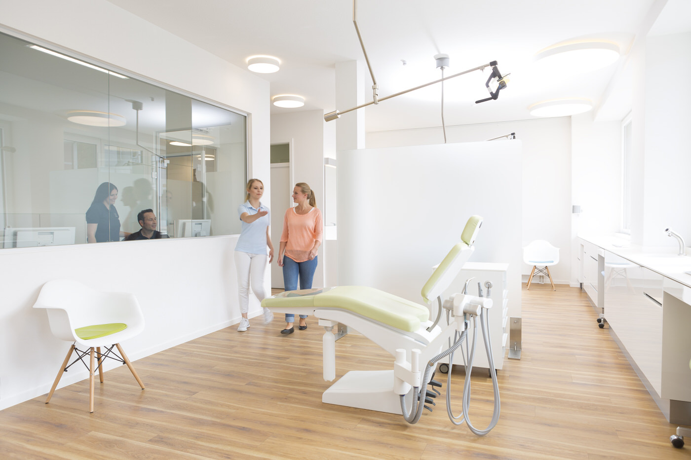 Moderne kieferorthopädische Behandlungszimmer mit hellem Interieur und Holzfußboden. Eine Mitarbeiterin erklärt einer Patientin die Umgebung, während im Hintergrund durch eine Glaswand das Team am Empfang sichtbar ist. Im Vordergrund ein Behandlungsstuhl mit medizinischer Ausstattung.