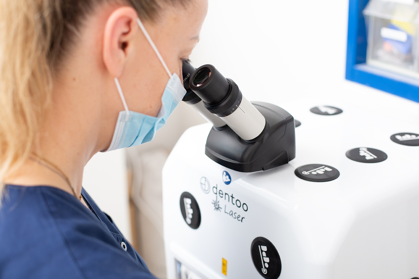 Eine medizinische Fachkraft mit Mundschutz blickt durch das Okular eines Mikroskops, das auf einem Dentallaser-Gerät mit der Aufschrift „dentoo Laser“ montiert ist. Das Bild vermittelt einen Eindruck von Präzision und moderner zahnmedizinischer Technik.